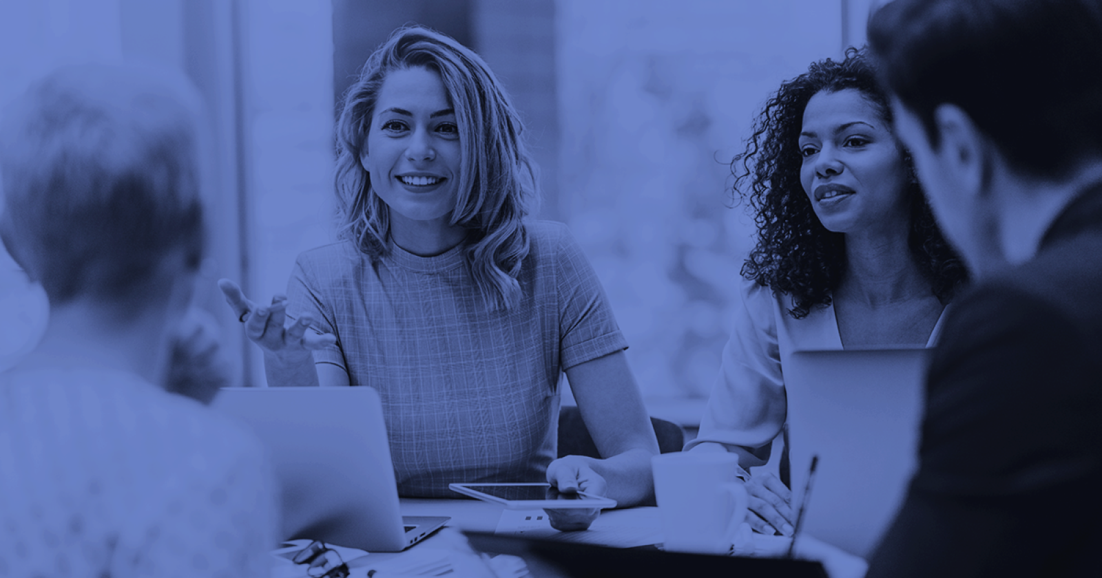 Group of people in a meeting, smiling and discussing. Laptops on the table. Blue tint.