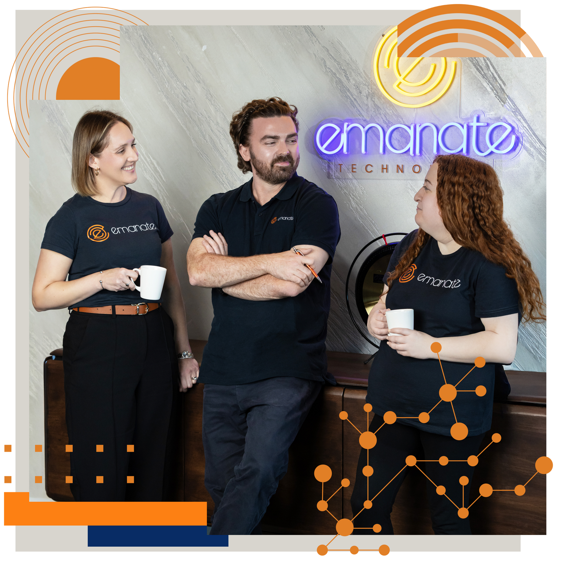 Three people in navy shirts with Emanate logo, standing in office; two holding mugs, smiling near