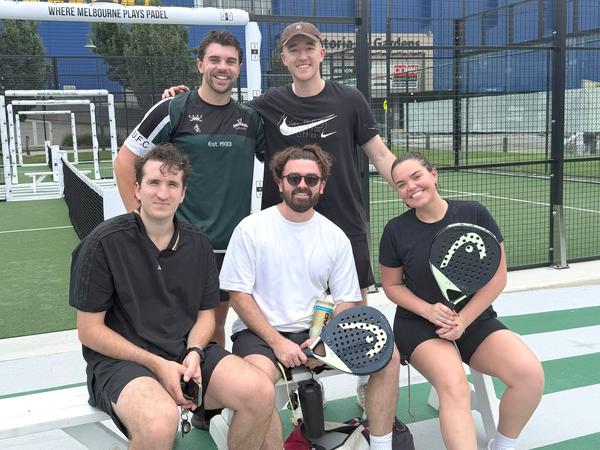 Five people sit on a bench on a padel court, some holding rackets, smiling after a game.