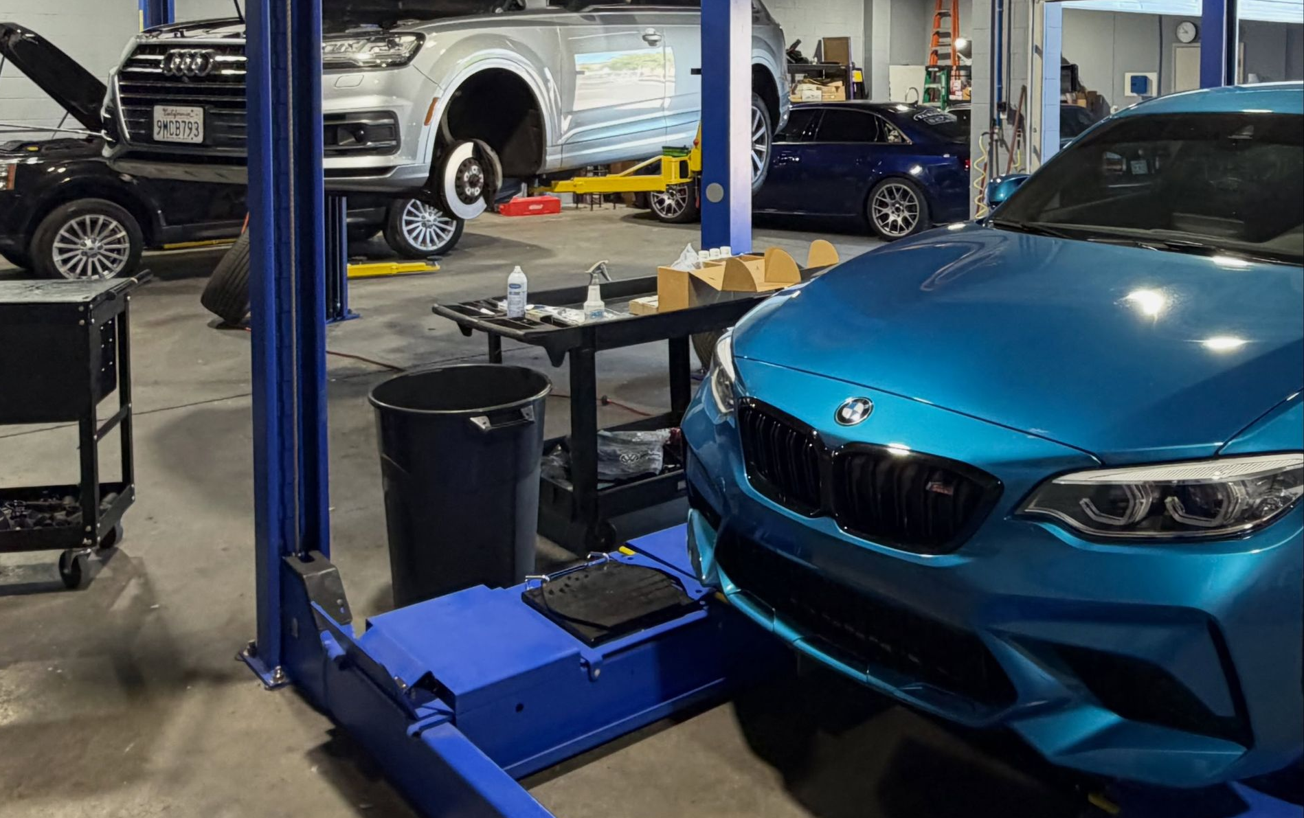 Blue sports car in auto repair shop beside a hydraulic lift, with another vehicle in the background.