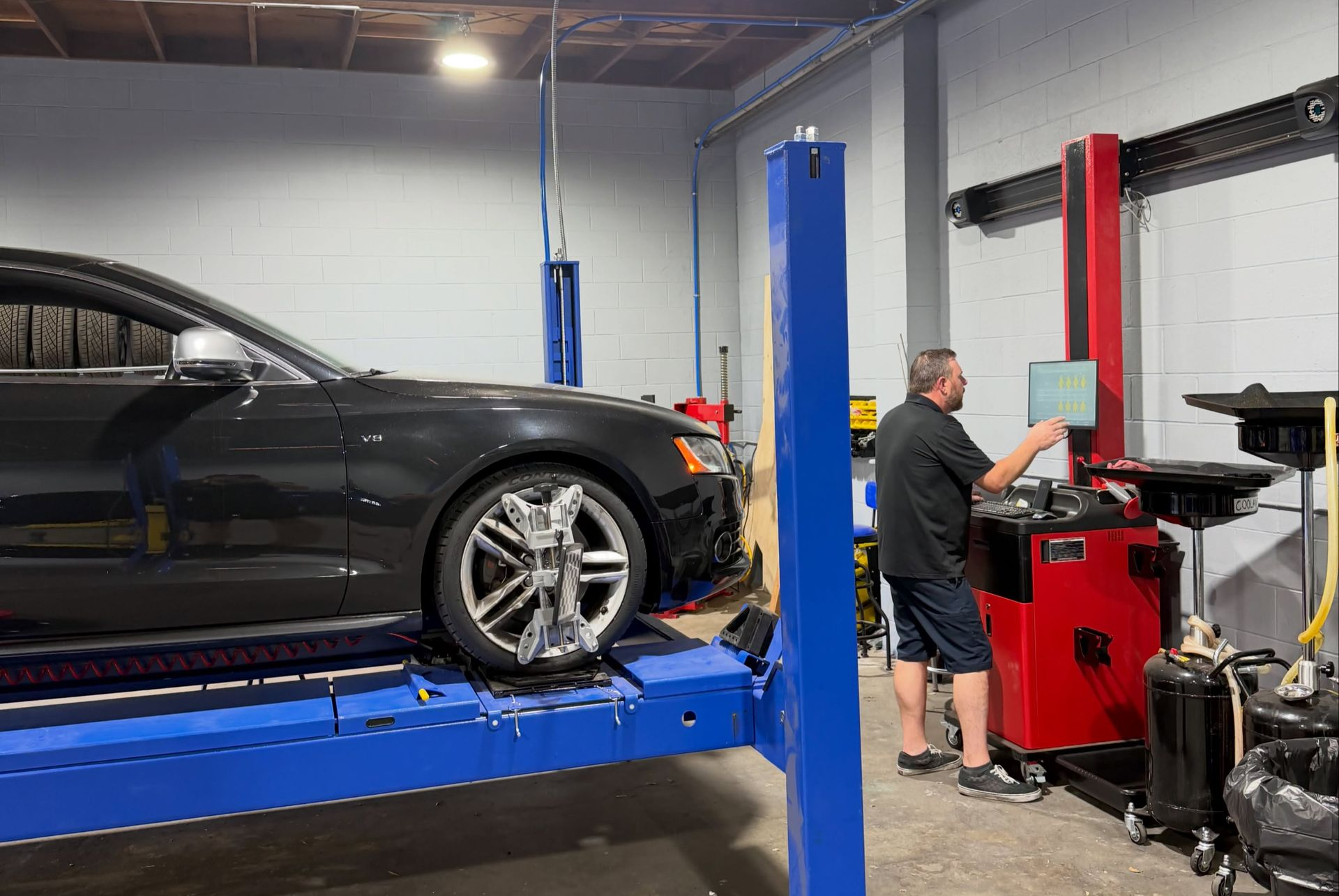 Black car on a blue lift in an auto shop while a mechanic works at a red machine nearby