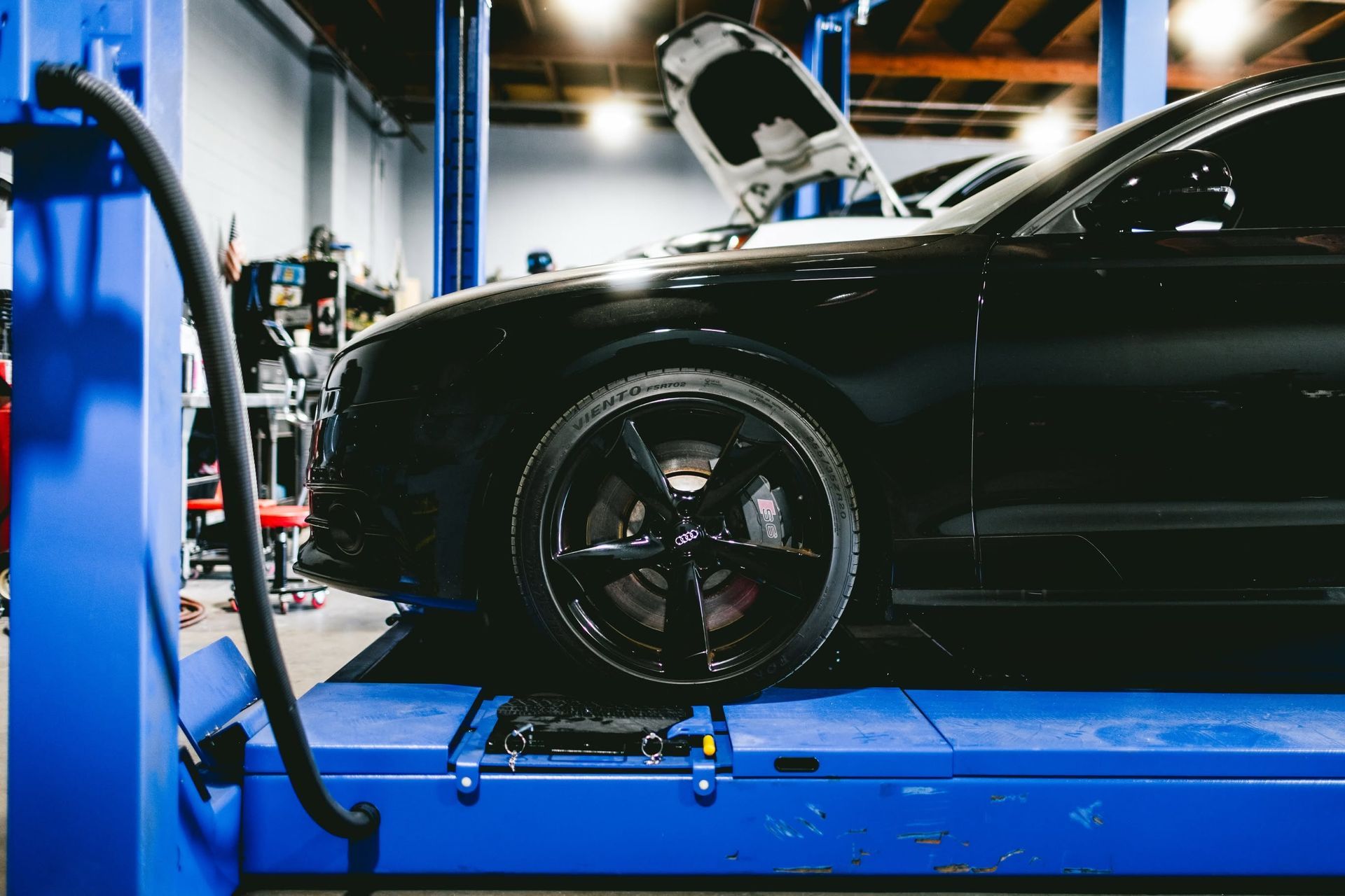 Black car on a blue lift in an auto repair shop, with wheels removed and hood open.