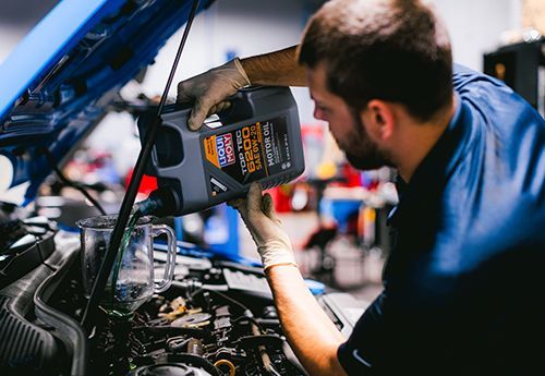Mechanic pouring oil into a car engine with the hood open in a garage