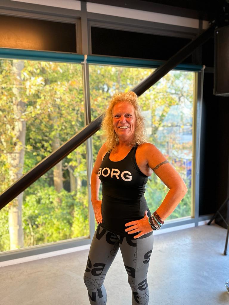 Een vrouw in een zwarte tanktop en grijze broek staat voor een raam.