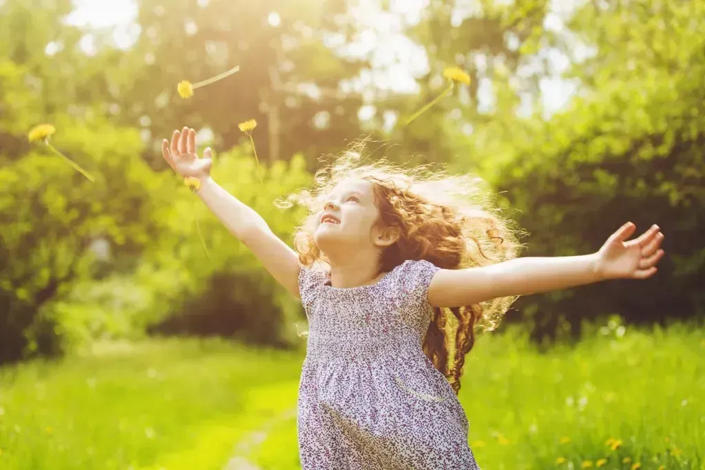 Child with arms outstretched in a sunlit green field, holding a flower and smiling upward