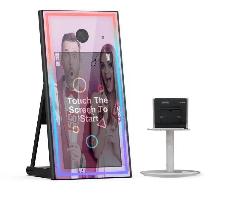 Photo booth mirror with a touch screen, next to a printer on a stand. Two people are reflected in the mirror.