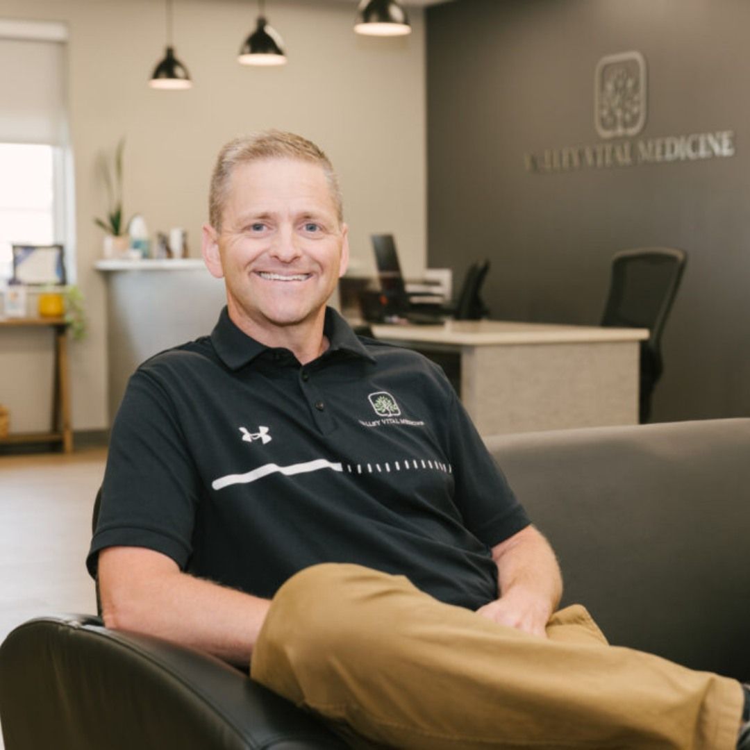 Smiling man seated in a medical clinic office with a reception desk and logo in the background