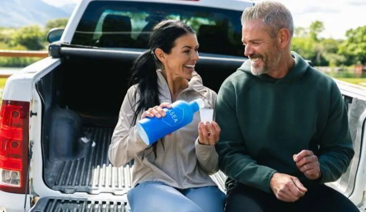 Two people smiling and sitting on the back of a pickup truck, sharing a snack outdoors.