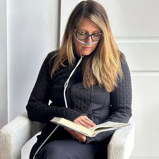 Woman in glasses reading a book in a white armchair by a bright wall