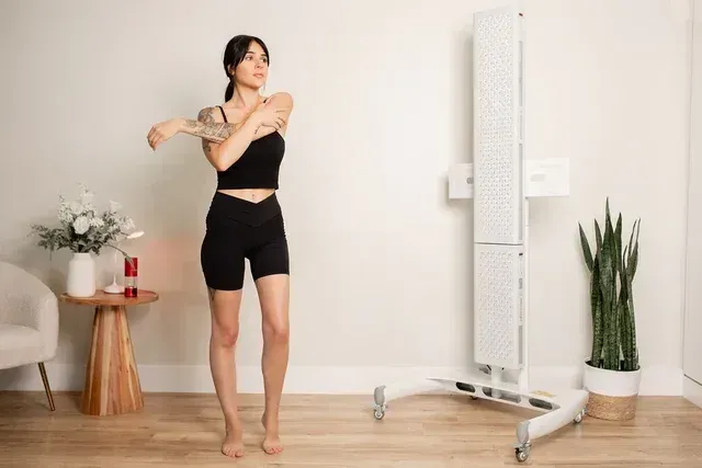 Woman stretching in a bright room beside a full-length mirror and potted plant.