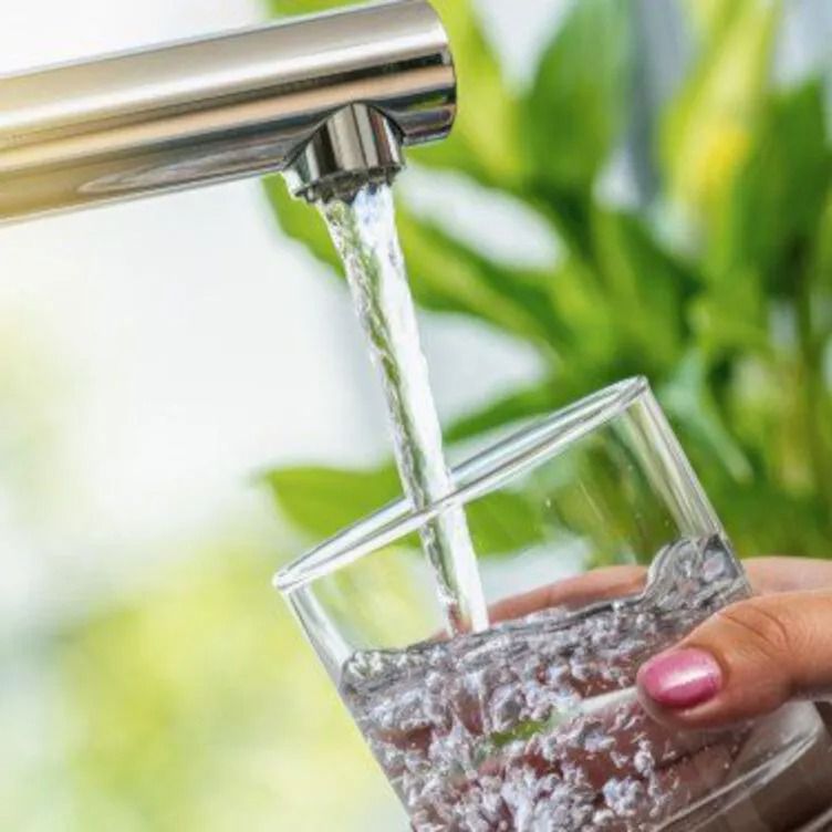 Water pouring from a tap into a clear glass held by a hand, with green plants in the background