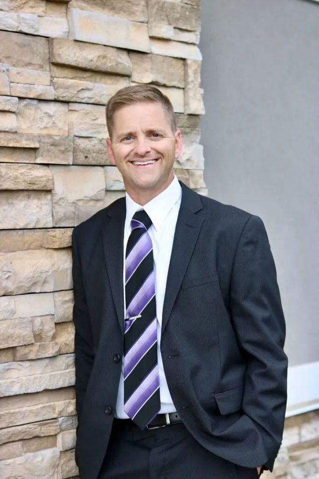 Smiling man in a dark suit and striped tie standing beside a stone wall