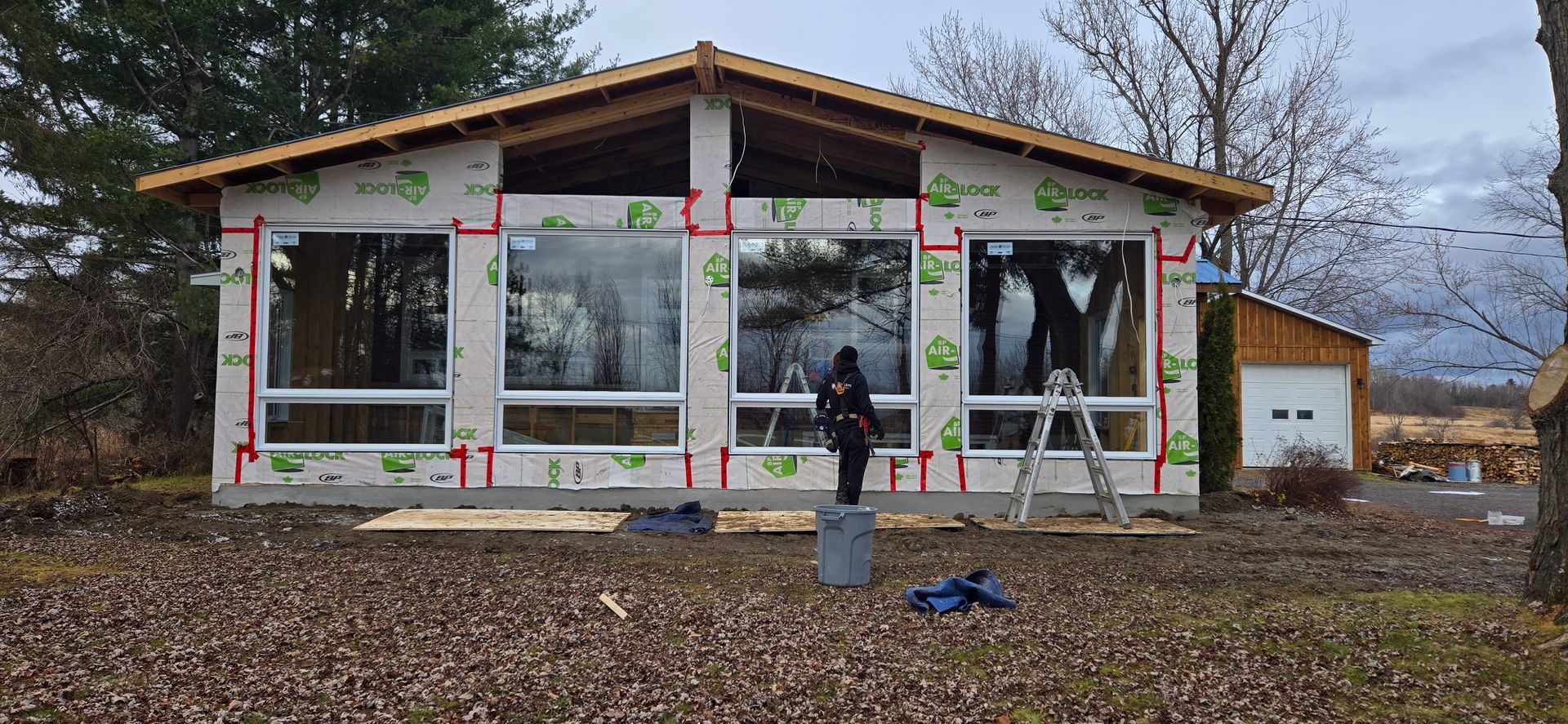 Maison partiellement rénovée, fenêtres scotchées, une échelle et un ouvrier devant, entourée de feuilles mortes.