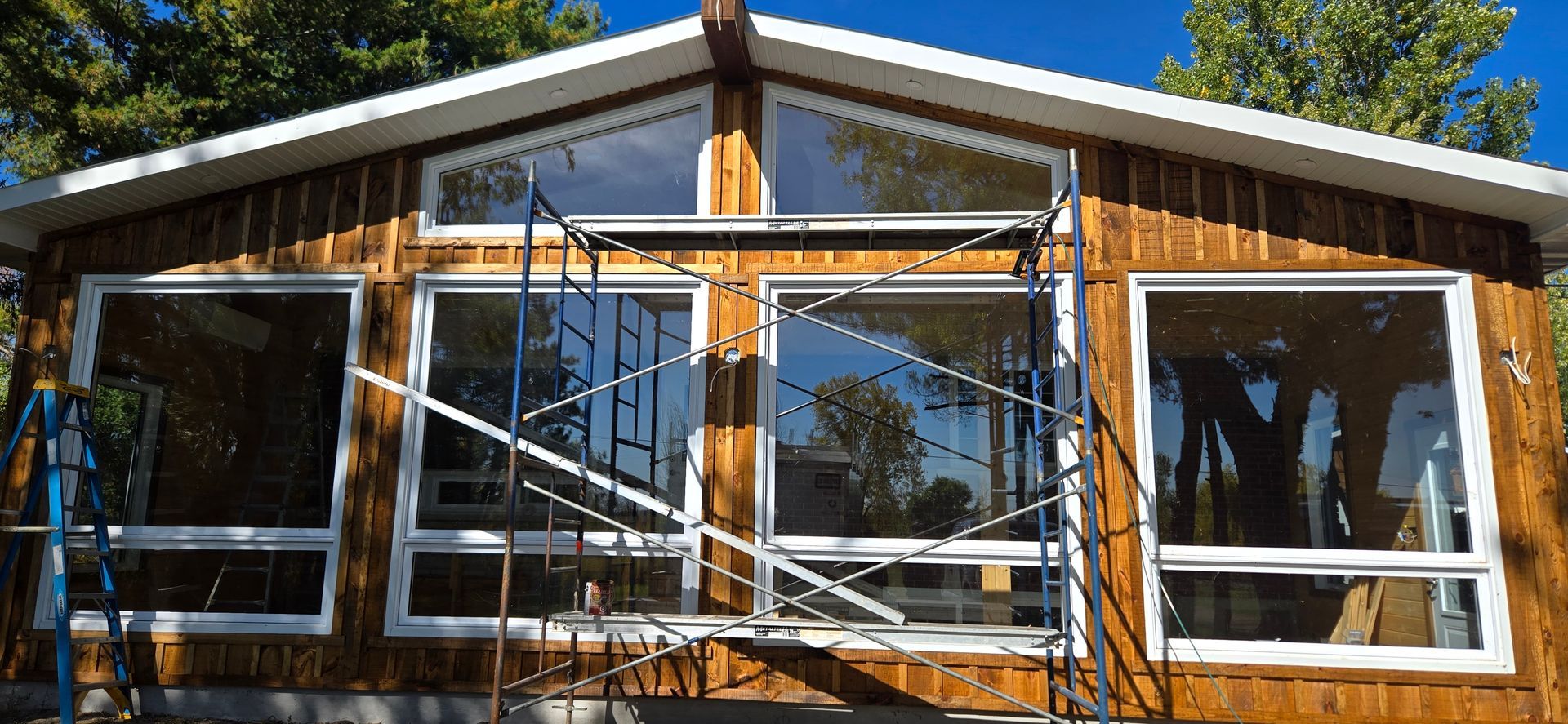 Façade de cabane en bois avec de grandes fenêtres et une porte vitrée centrale baignée de soleil