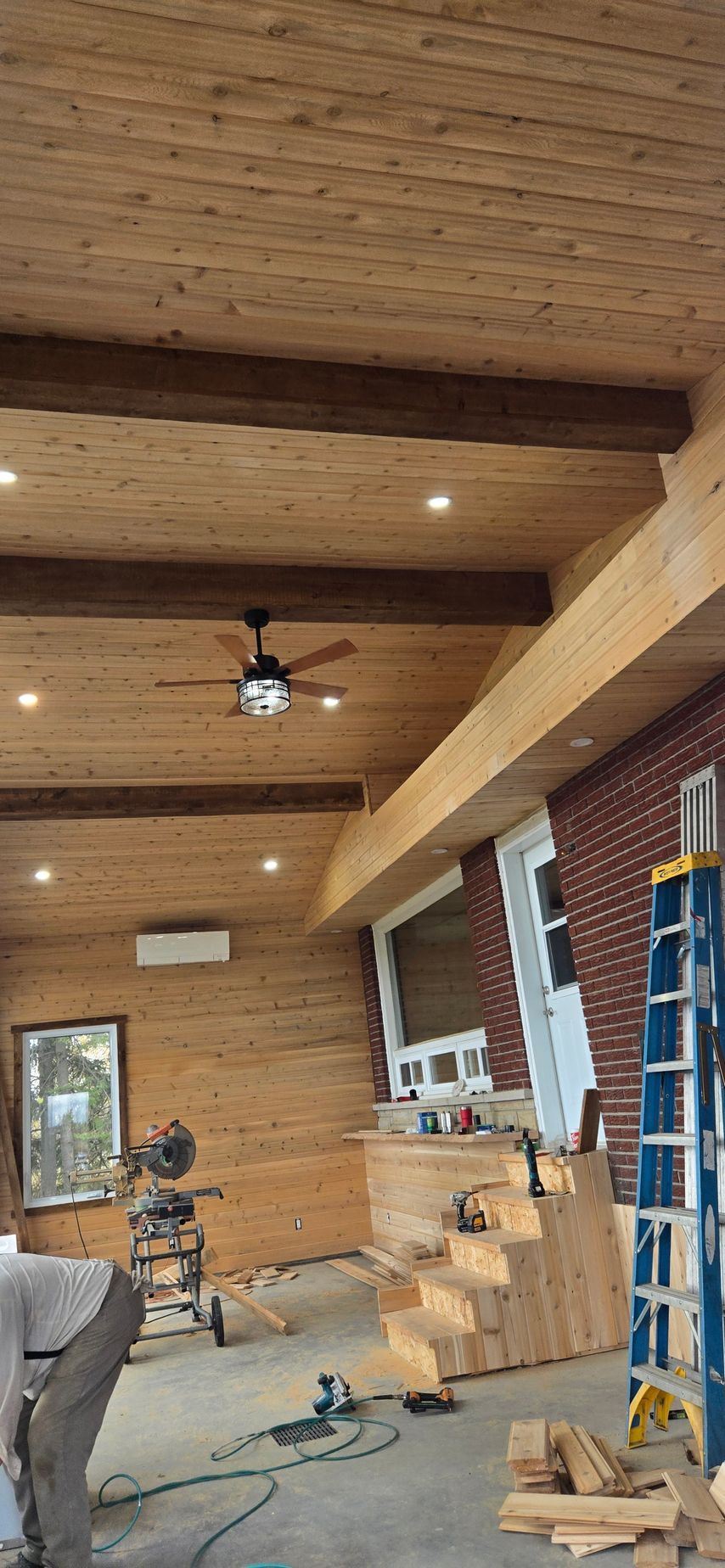 Intérieur en bois en cours de construction, avec poutres apparentes, échelle, outils et planches empilées contre un mur de briques.