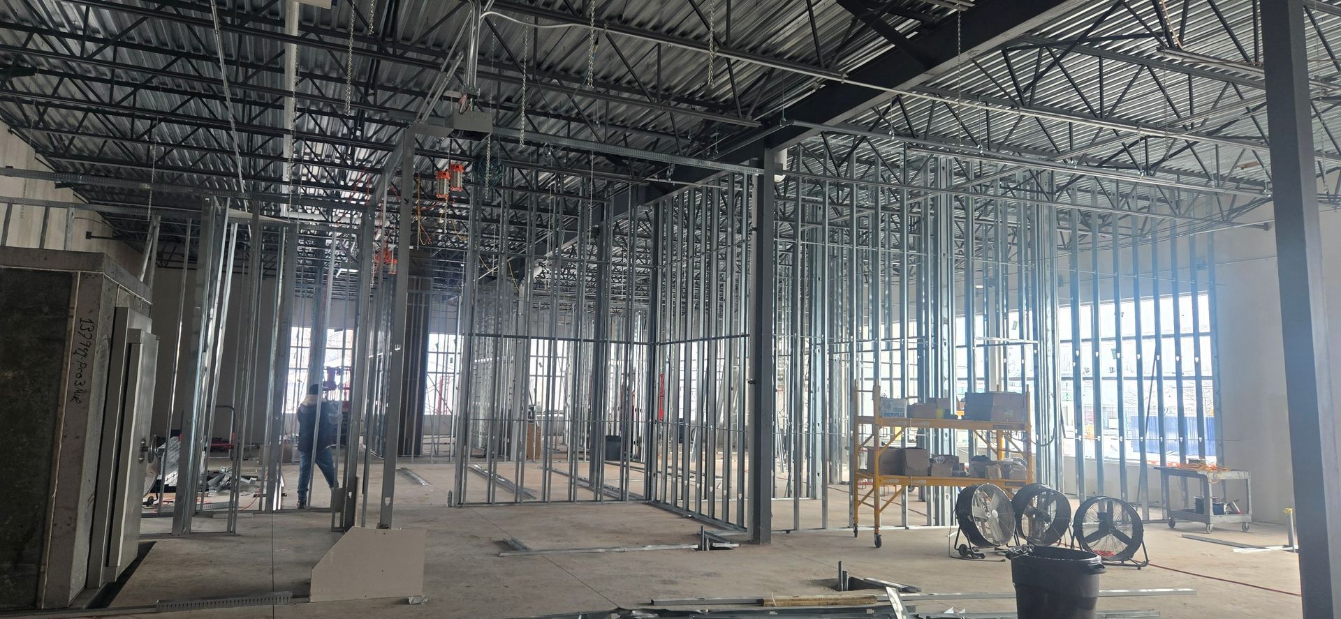 Grand intérieur inachevé avec charpente métallique apparente, ouvriers et engins de chantier sur un sol en béton.