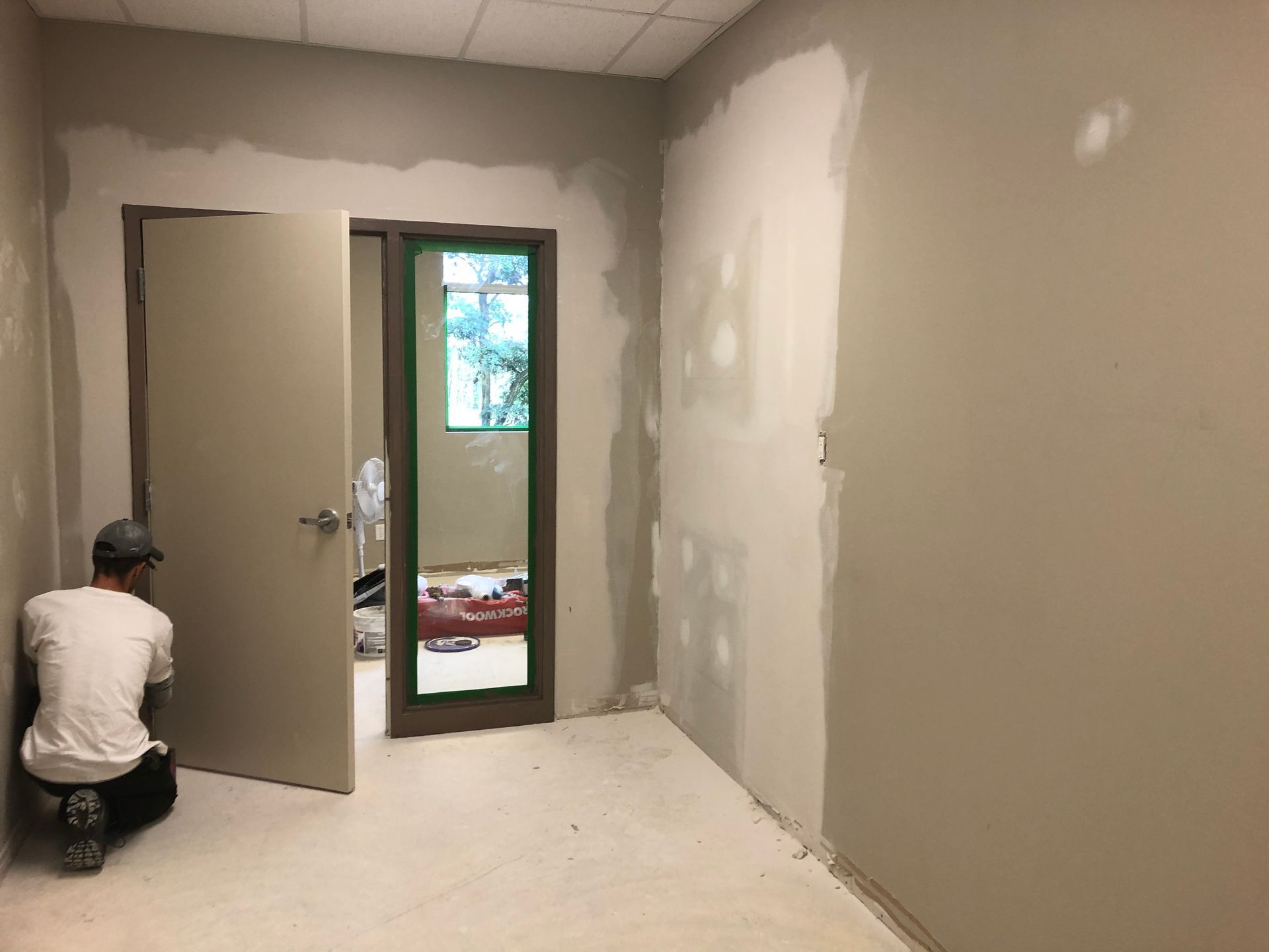 Un ouvrier agenouillé dans une pièce en rénovation, aux murs gris et à la porte ouverte donnant sur un couloir lumineux.