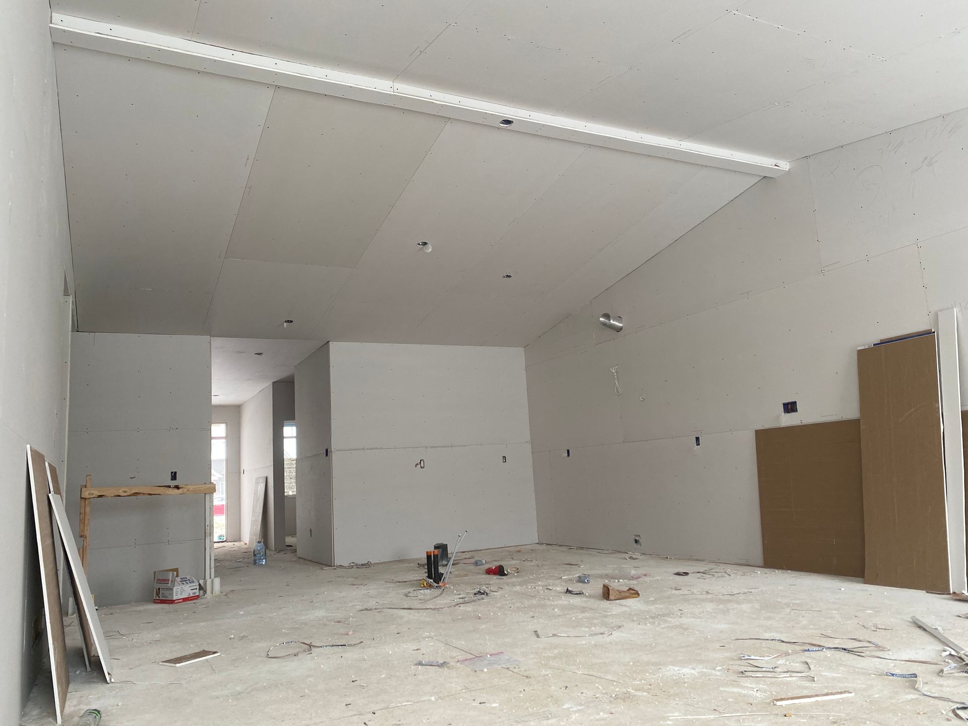 Pièce vide et inachevée, aux murs blancs, au plafond apparent, jonchée de débris de construction et avec une porte ouverte.