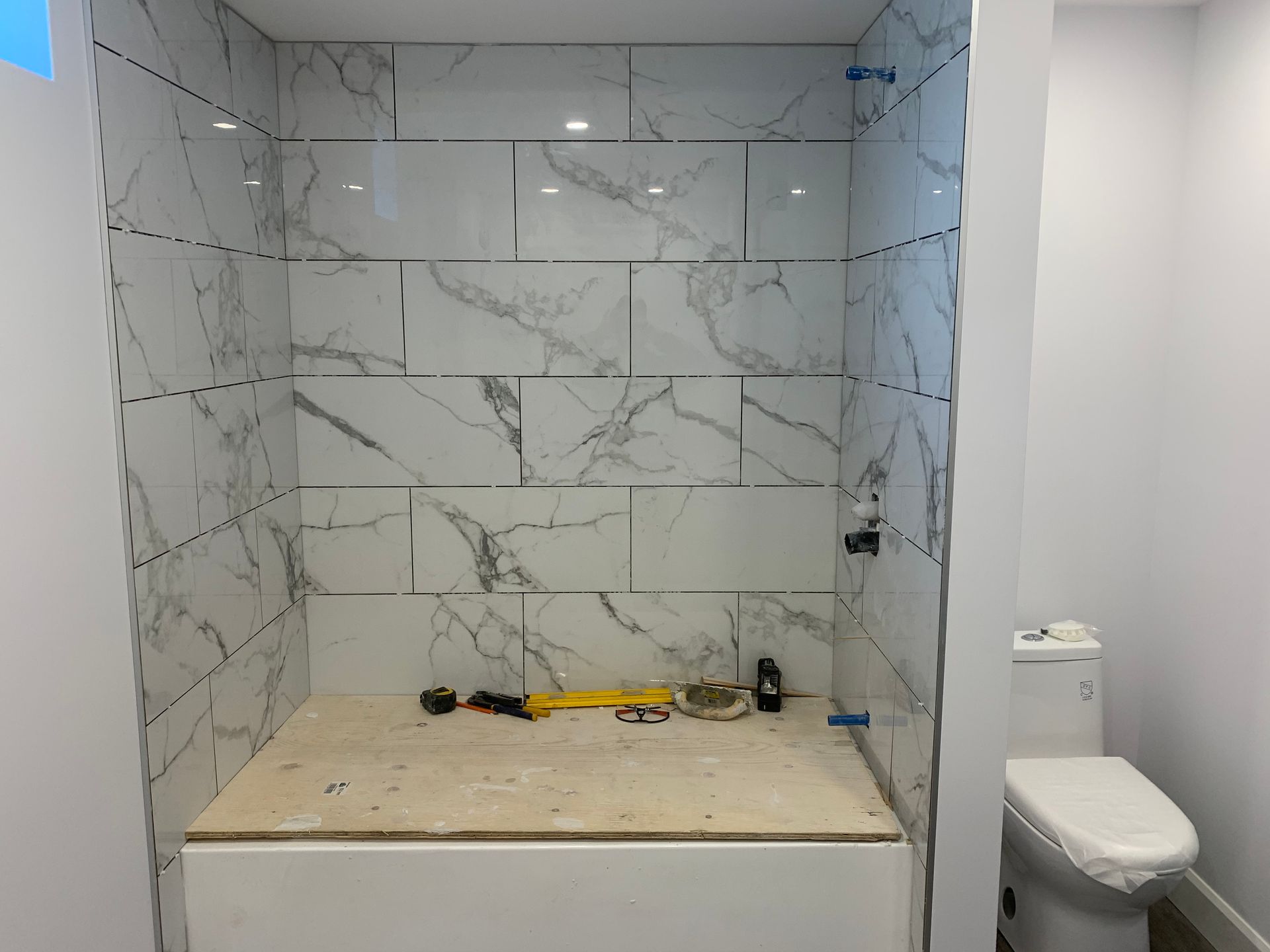 Alcôve de douche partiellement carrelée dans une salle de bain en construction, avec des outils sur un établi rudimentaire et des toilettes à côté.