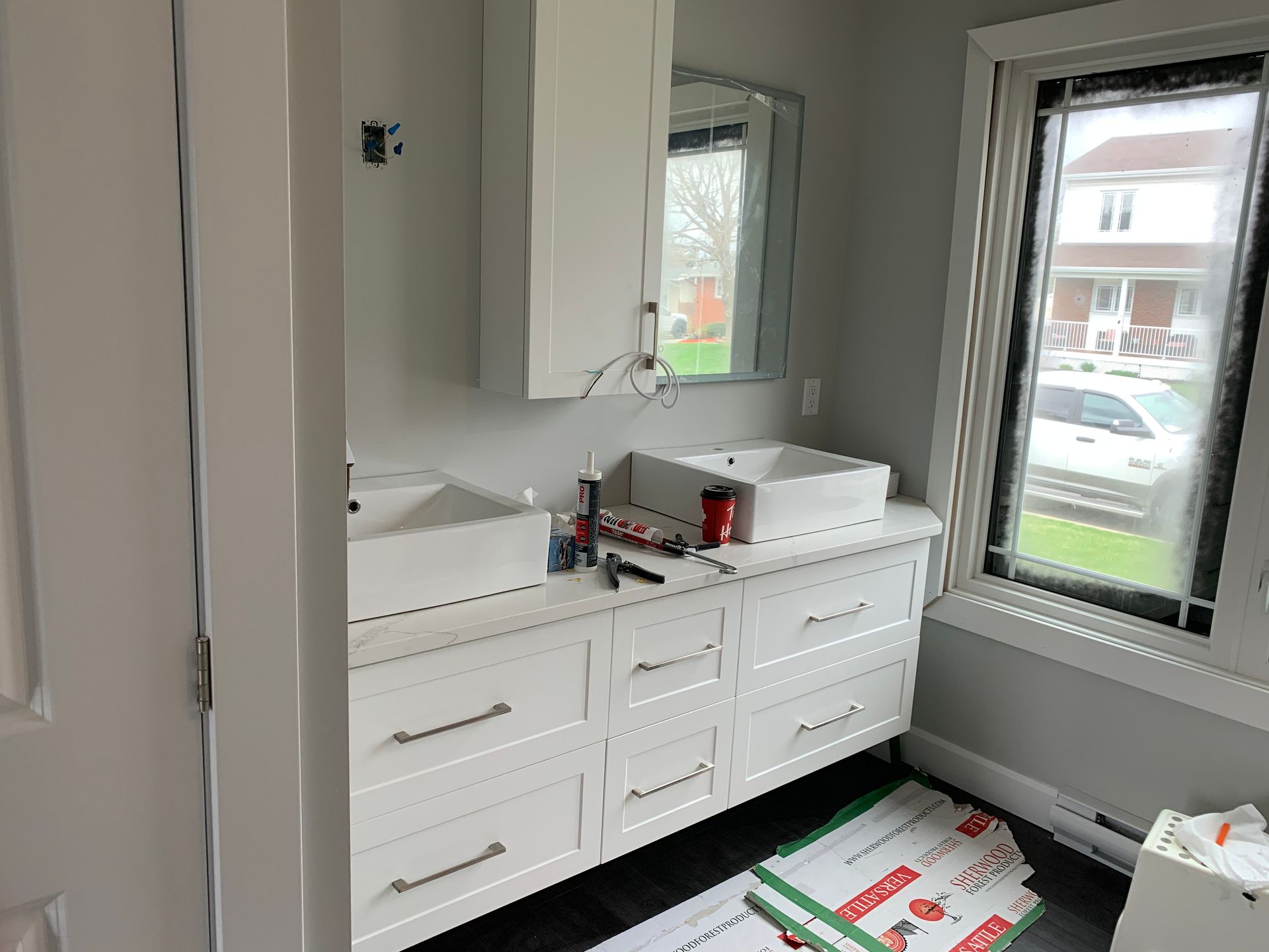 Meuble-lavabo blanc avec miroir et articles de toilette, à côté d'une fenêtre et d'une boîte ouverte posée au sol.