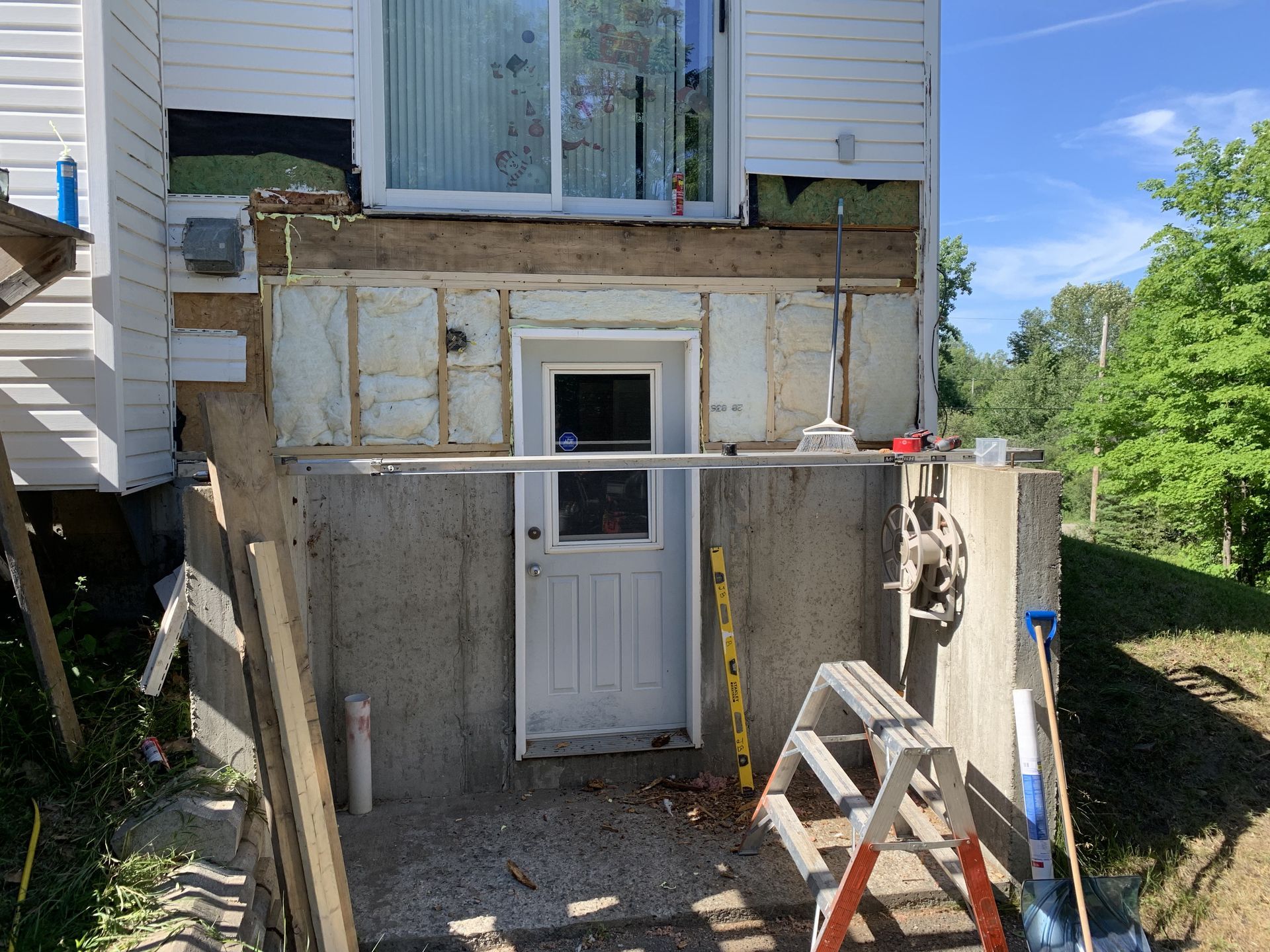 Façade de maison partiellement rénovée avec une porte blanche, isolation apparente et une petite échelle près des fondations