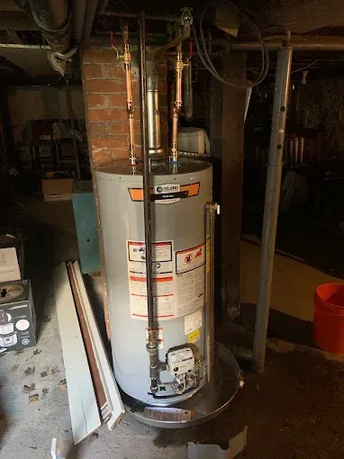 Water heater in a basement utility area with exposed pipes and unfinished brick walls
