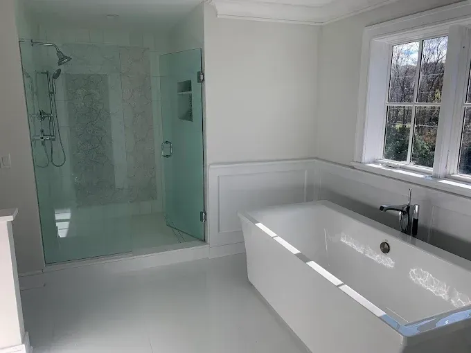Accessible white bathroom with walk-in glass shower, bathtub, and window