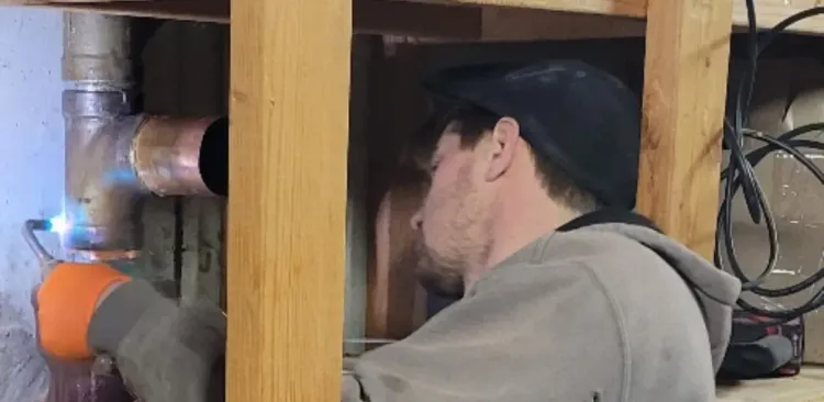 Person working under a sink with exposed pipes and orange insulation in a crawlspace.