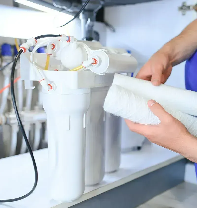 Hand holding a white water filter cartridge beside a countertop filtration system with clear canisters
