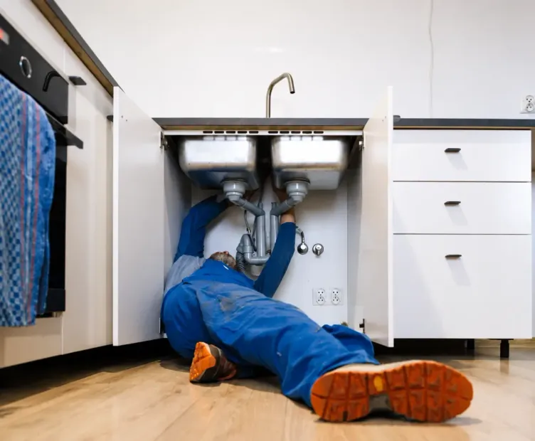 Plumber in blue overalls repairing a sink under a bathroom cabinet, tools nearby.