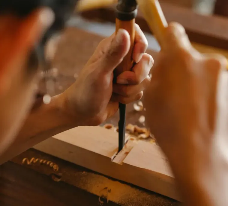 Hands use a chisel and mallet to carve wood on a wooden workbench, creating a mortise joint.