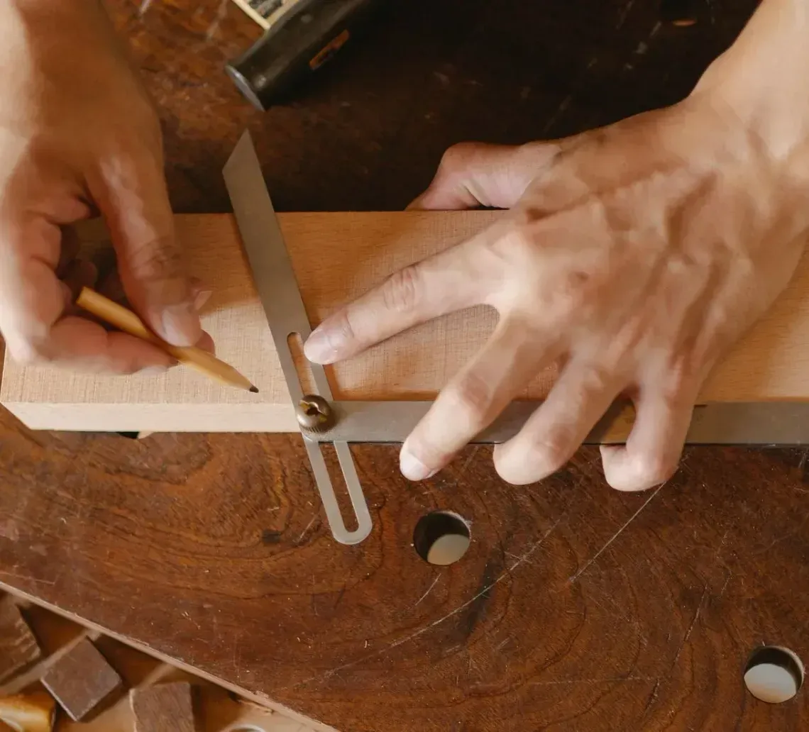 Person using a combination square to mark a line on a wooden plank with a pencil, on a wooden workbench.