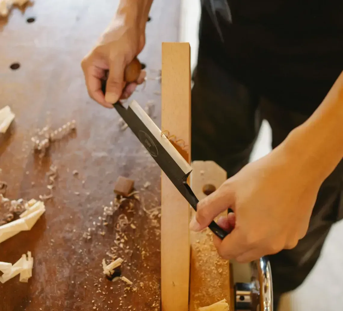 Person using a drawknife to shape a wooden board on a workbench, wood shavings visible.