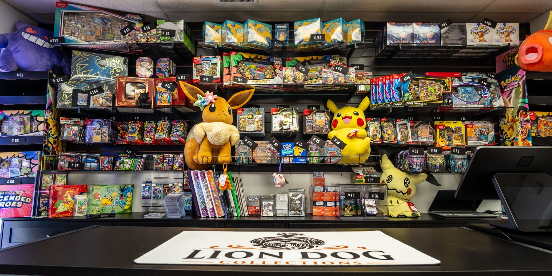 Pokémon card shop display with plush toys, booster packs, and a laptop on the counter