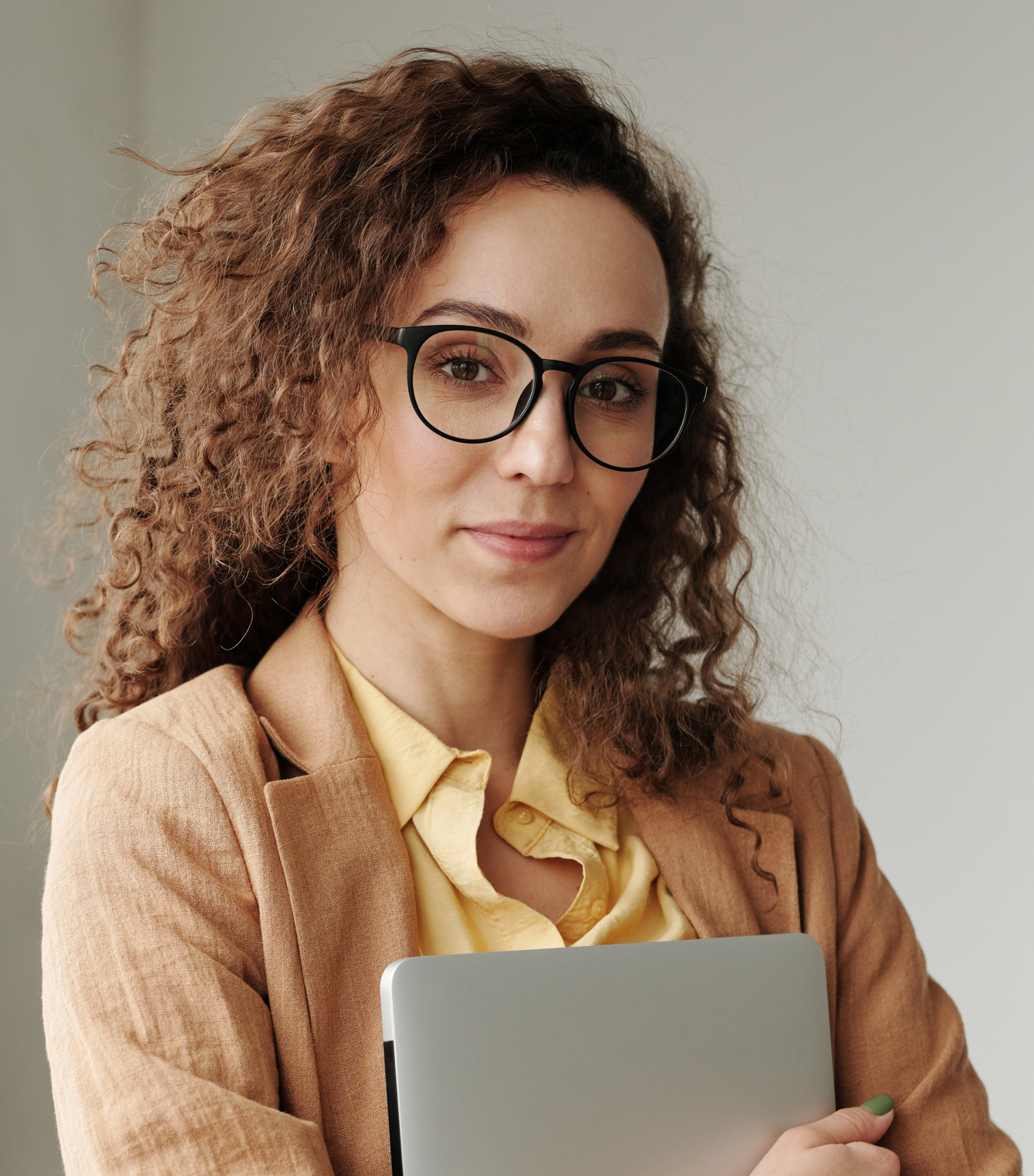 Woman with curly hair and glasses holds a laptop, wearing a tan blazer and yellow shirt.