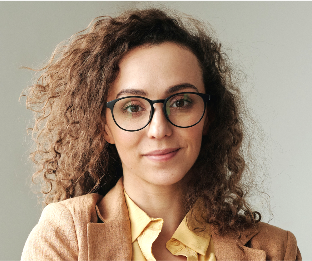A woman with curly hair wearing glasses and a tan jacket.