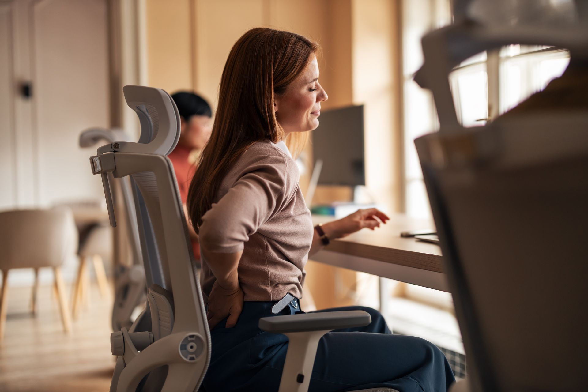 Woman at desk with back pain, hand on lower back, office setting.