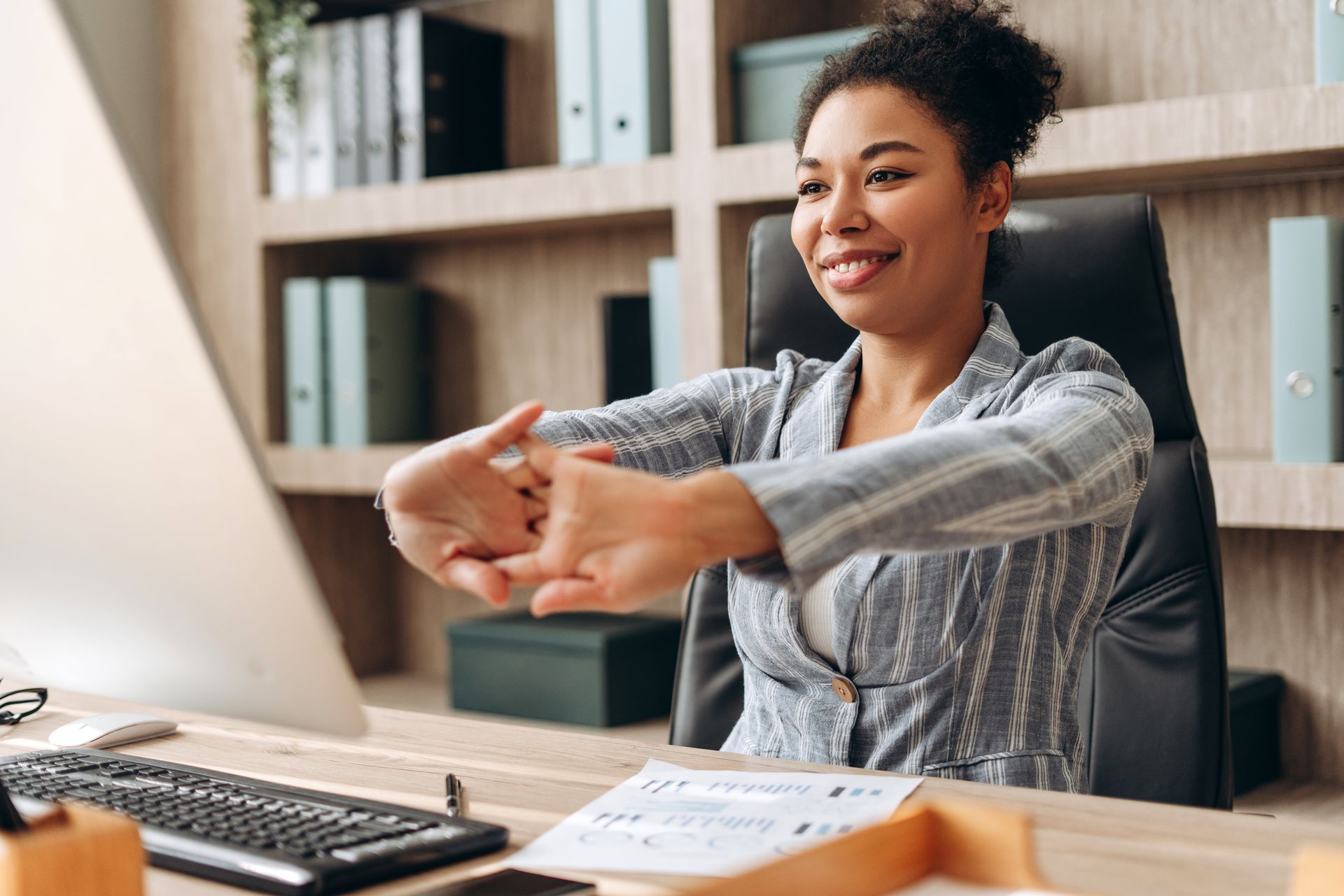 Woman stretching at a desk in a bright office, smiling near a laptop and keyboard.