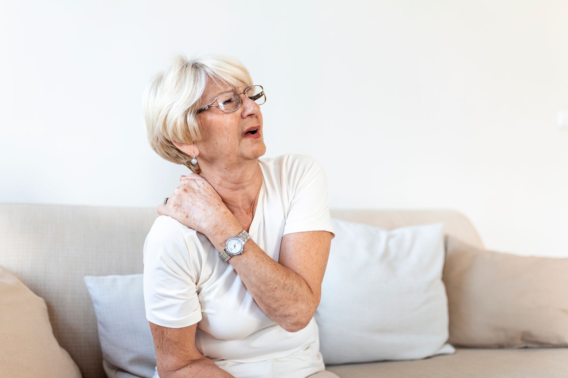 Elderly woman in a white shirt sitting on a couch, touching her neck with a pained expression
