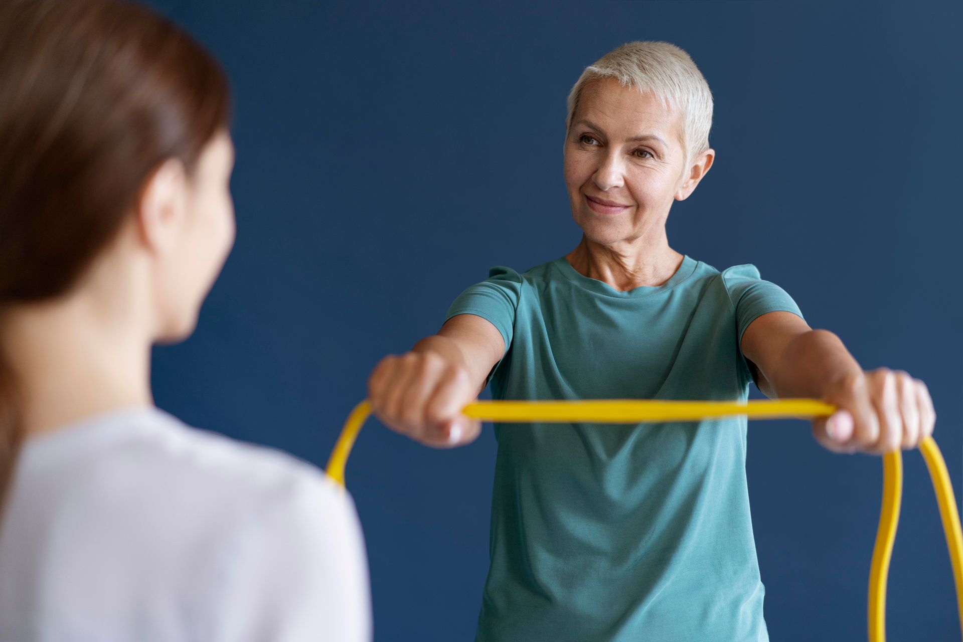 Woman exercising with resistance band, guided by another person. Teal shirt, blue background.