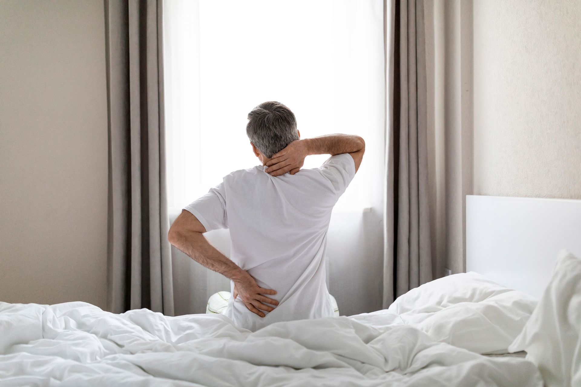 Person sitting on a bed, holding their lower back, suggesting back pain in a bright bedroom.