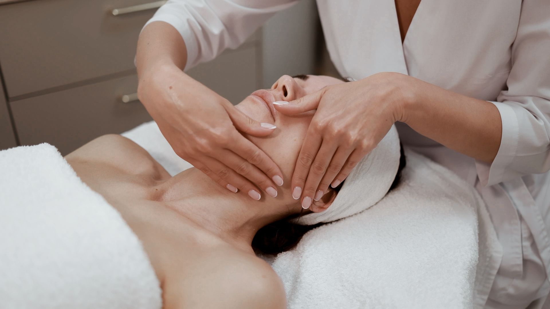 Woman receiving a facial massage, lying down with eyes closed, hands on cheeks, spa setting.