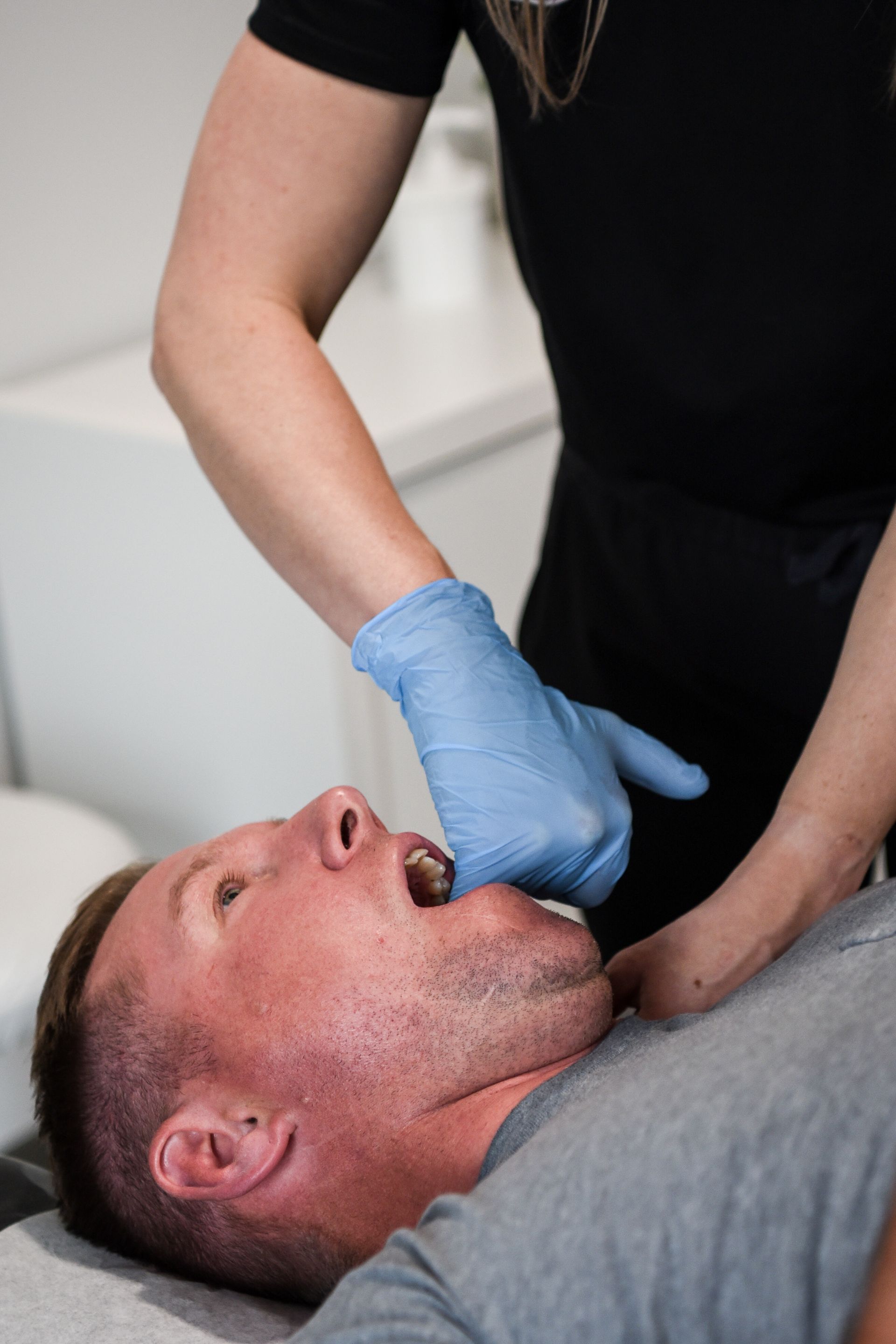 Person receiving jaw massage; gloved hand inside mouth, other hand on chin, indoors.