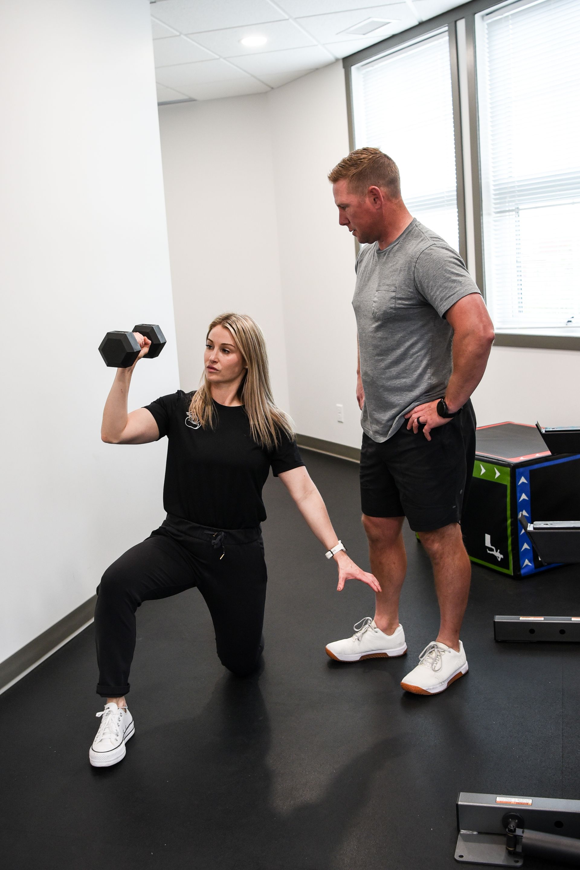Woman doing a dumbbell exercise while kneeling, watched by a trainer in a gym.