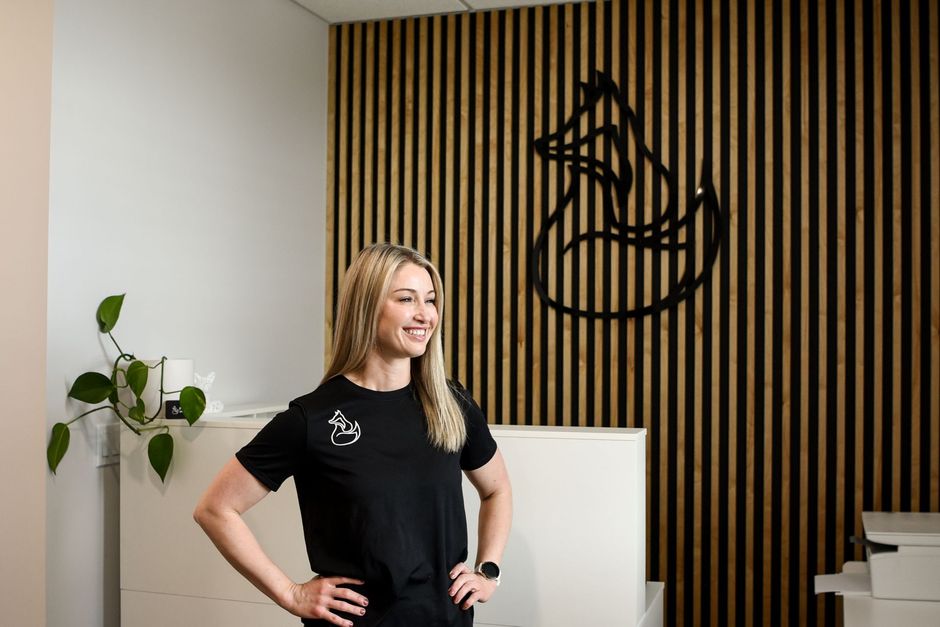 Woman in black t-shirt smiles, hands on hips, in front of wooden paneling with fox logo.