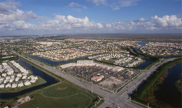 Aerial view of a suburban neighborhood with canals, roads, and water under a partly cloudy sky