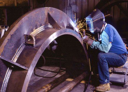 Welder in blue protective gear welding a large metal pipe in an industrial workshop