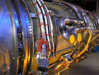 Close-up of a large metallic engine with pipes, cables, and glowing yellow lights in an industrial setting