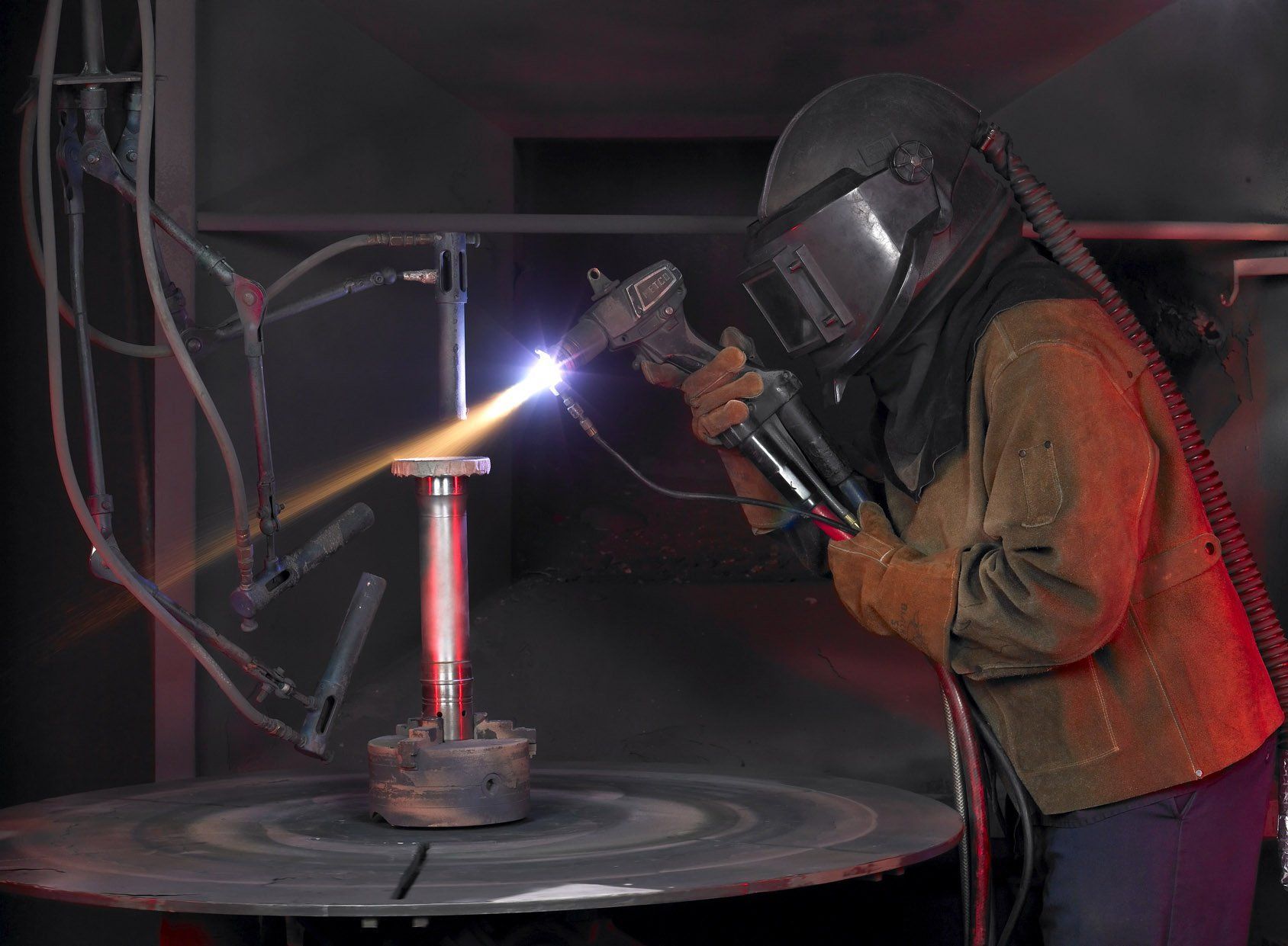 Person in a welding mask and brown jacket welding a red metal cylinder on a workbench in a dark workshop.