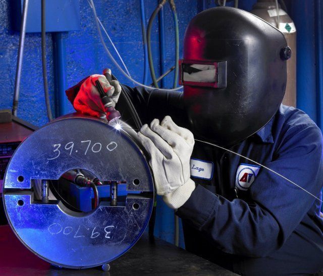 Welder in black helmet and gloves inspecting a large metal component marked with numbers and weld seams.